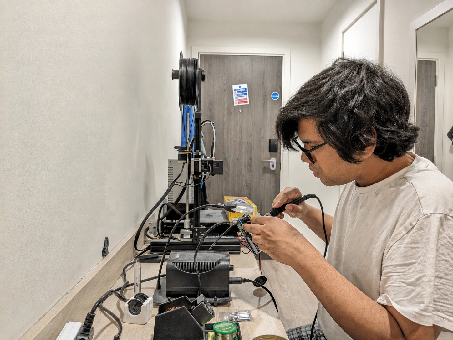 Alok soldering electronics beside a 3D printer in his workshop